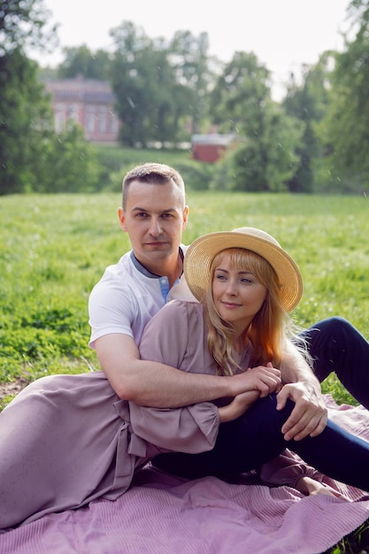 Man and a woman in love in a dress and hat are sitting on a blanket on a green field under a tree in summer