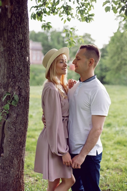 Man and a woman in love in a dress and hat are stand on a green field under a tree in summer