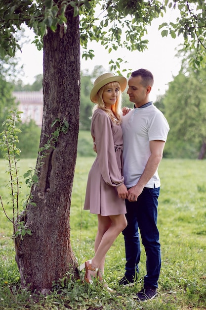 Man and a woman in love in a dress and hat are stand on a green field under a tree in summer