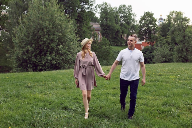 Man and a woman in love in a dress and hat are walk on a green field under a tree in summer