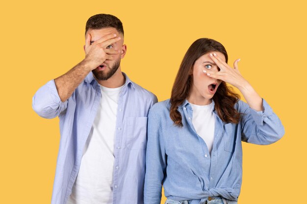 Photo man and woman peeking through fingers showing playful fear on yellow background