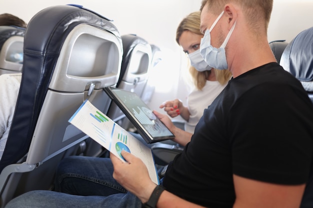 Photo man and woman in protective medical masks flying in airplane and discussing diagram on documents