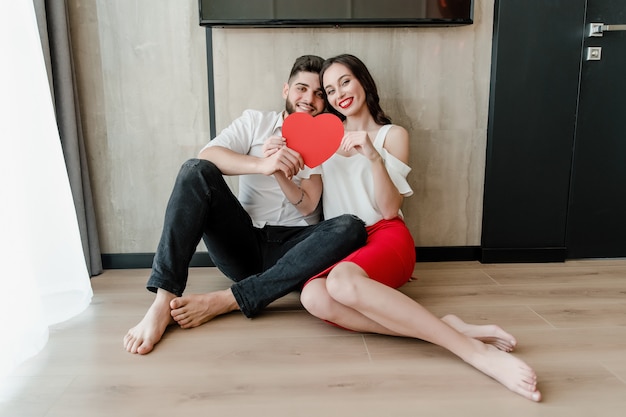 Man and woman behind red heart shaped valentine card sit at home on the floor