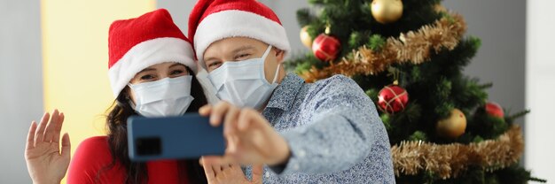 Man and woman in santa hats and protective masks on faces are holding a mobile phone and waving near