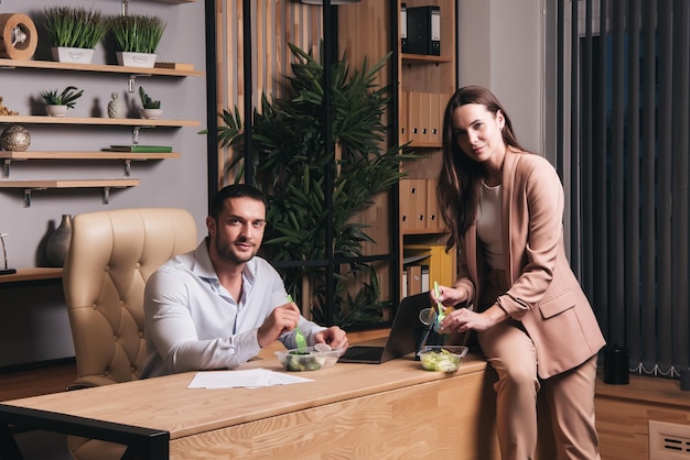 A man and woman sit at a desk with a bowl of salad.