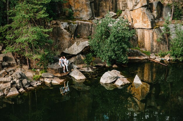 Photo man and a woman sit on the rocks, under the tree, near the forest and the lake
