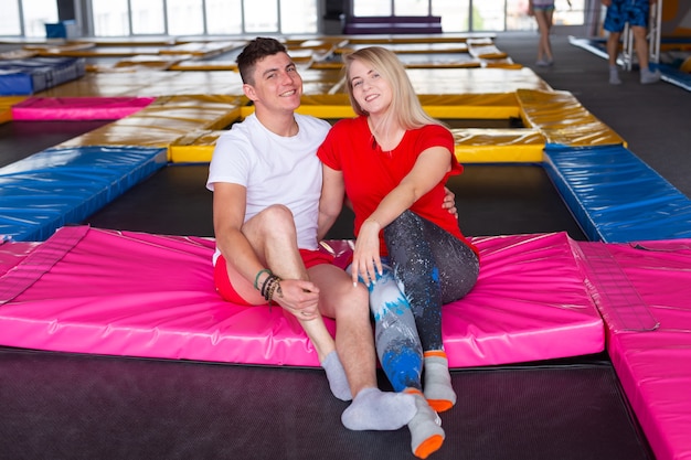 Man and woman sits together on a trampoline indoors.