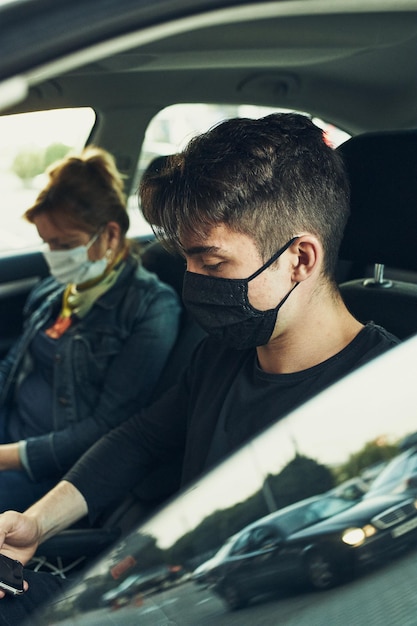man and woman sitting in a car using smartphones wearing the face masks to avoid virus