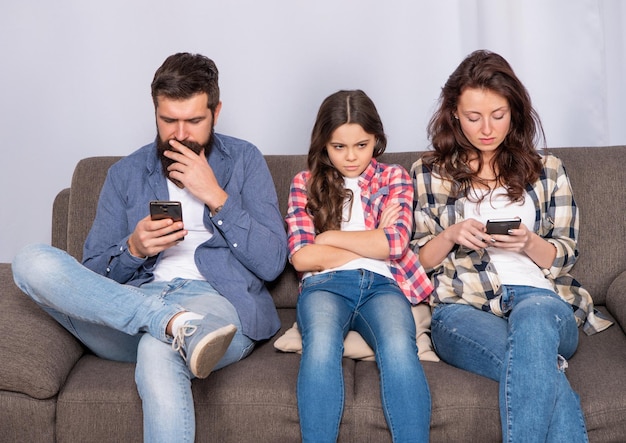 Man and woman sitting together on sofa engaging with smartphones ignoring each other and unhappy girl child at home, family.