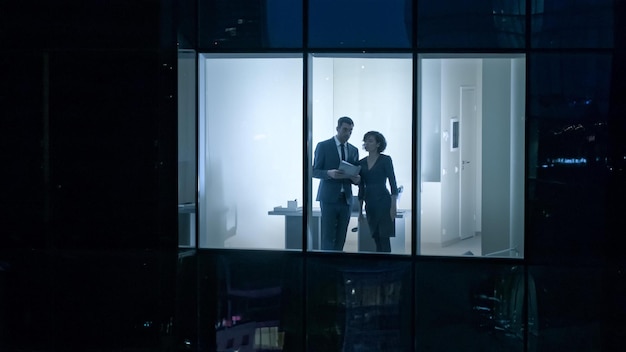 A man and woman stand in a window looking out at night.