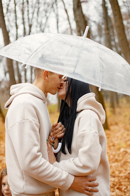 Man and woman standing in autumn forest and kissing under transparent umbrella Brunette man and woman falling in love Family wearing beige sportive costumes