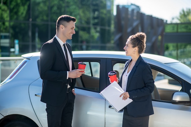 Man and woman standing near car with coffee in hand.