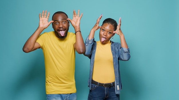 Man and woman sticking tongue out and fooling around in front of camera. Playful people doing funny and comic faces, enjoying goofy joke together in studio. Couple showing childish behavior.