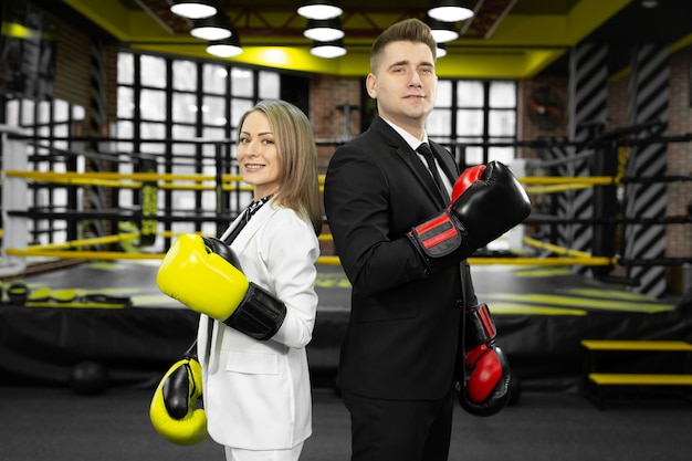 Man and woman in a suit and boxing gloves stand in front of the ring with their backs to each other.