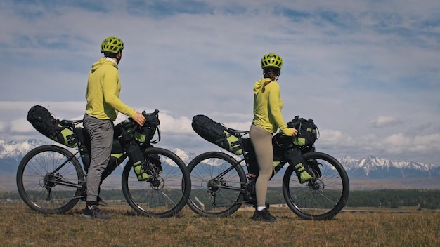 The man and woman travel on mixed terrain cycle touring with bikepacking. The two people journey with bicycle bags. Mountain snow capped.