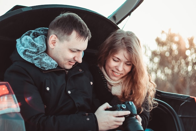 A man and a woman on truck of the car. A happy couple, wrapped in a warm knitted blanket, watching photos on camera against the backdrop of the winter forest,