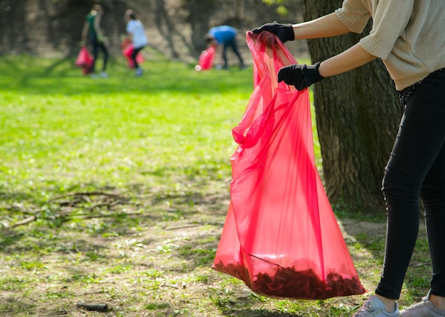 Man and woman volunteer wearing picking up trash and plastic waste in public park. Young people wearing gloves and putting litter into red plastic bags outdoors