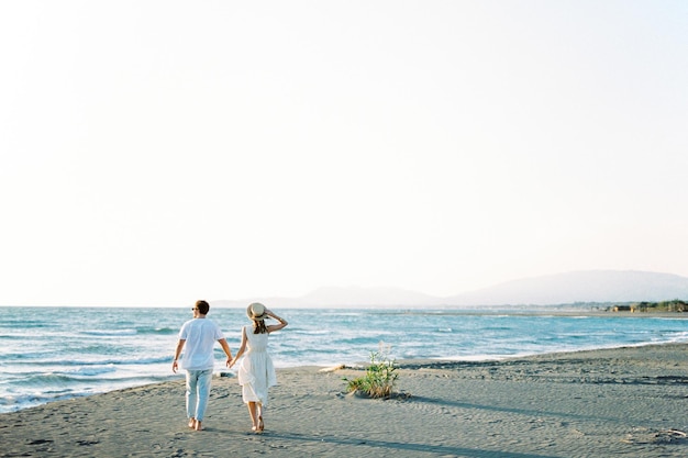 Man and woman walk holding hands on the sandy beach to the sea Back view