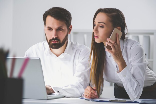 Man and woman in white clothes are working in an office together. He is typing and looking at his laptop screen. She is on her phone and reading.