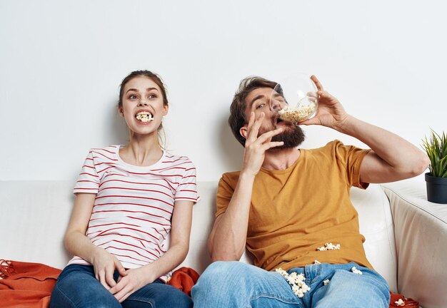 Foto un uomo e una donna su un divano bianco in una stanza luminosa un piatto di popcorn e amici un fiore in un vaso foto di alta qualità