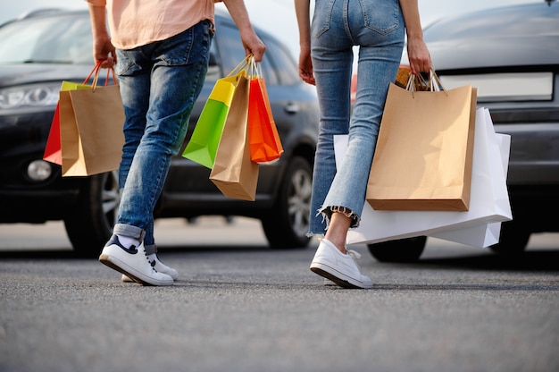 Uomo e donna con sacchetti di cartone sul parcheggio del supermercato. clienti felici che trasportano acquisti dal centro commerciale, veicoli