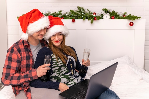 A man and a woman with champagne communicate online on a laptop. Couple drinking champagne in santa hats