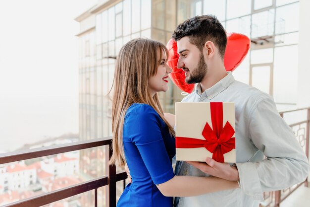 Мужчина и женщина с подарком и красные шары в форме сердца на балконе дома