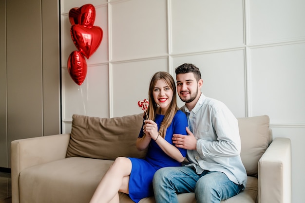 Man and woman with lollypop candy at home on sofa with heart shaped balloons