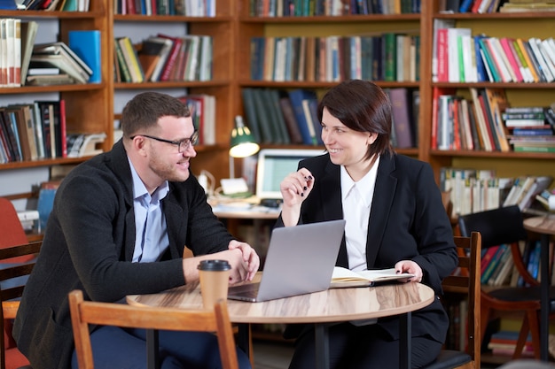 Uomo e donna che lavorano con il computer portatile che si siede nella stanza alla moda moderna con gli scaffali per libri in biblioteca o libreria