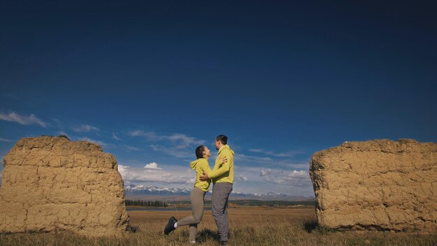 Uomo e donna in abbigliamento sportivo giallo verde. una bella coppia di viaggiatori si abbraccia e si bacia vicino alla vecchia pietra godendosi il paesaggio dell'altopiano. due viaggiatori stanno camminando sullo sfondo di montagne innevate.