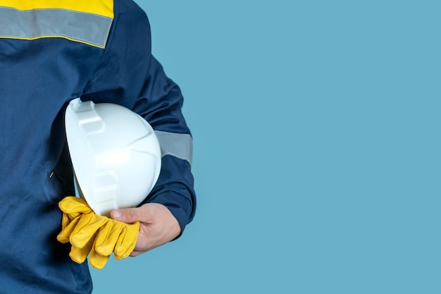 A man in work clothes holds a white protective helmet and gloves on a blue background in his hand. Free space for the inscription.