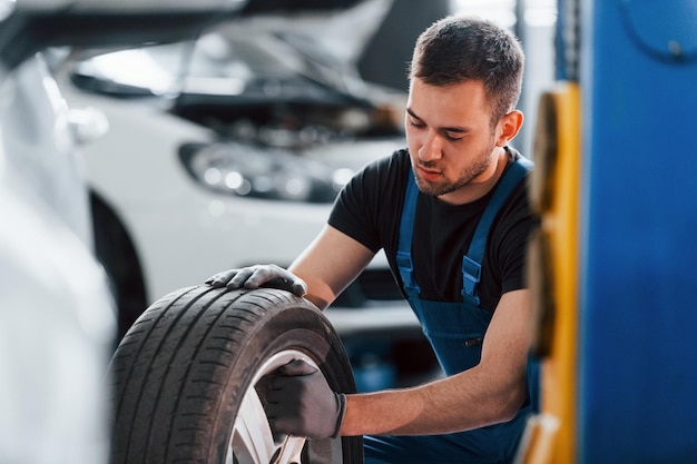 Man in work uniform sitting and changing car wheel indoors conception of automobile service