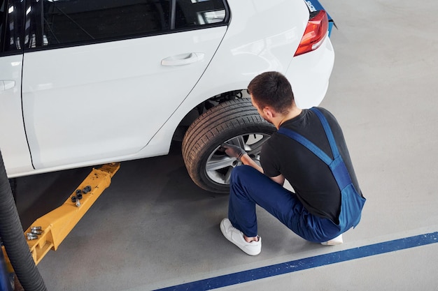 Man in work uniform sitting and changing car wheel indoors Conception of automobile service