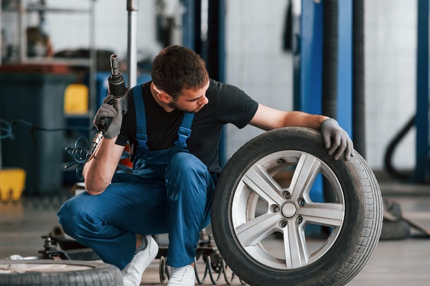 Man in work uniform sitting with car wheel indoors Conception of automobile service