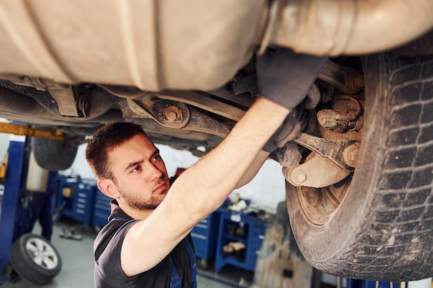 Man in work uniform standing under car and repairs it indoors Conception of automobile service