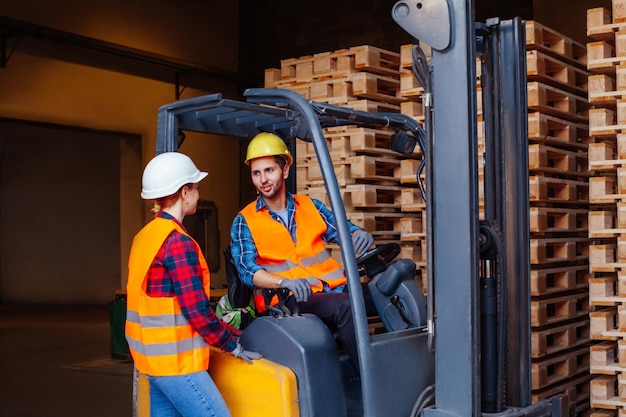 Man worker sitting in industrial forklift at warehouse talking to woman coworker