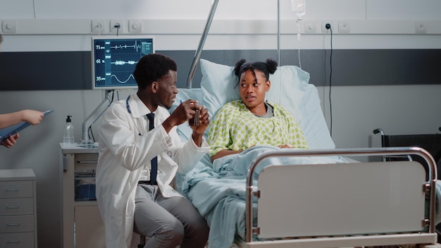 Man working as medic giving bottle of pills to ill woman in bed to cure sickness. Medical physician explaining prescription treatment and medicine in flask while sitting in hospital ward