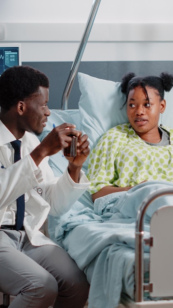 Man working as medic giving bottle of pills to ill woman in bed to cure sickness. Medical physician explaining prescription treatment and medicine in flask while sitting in hospital ward