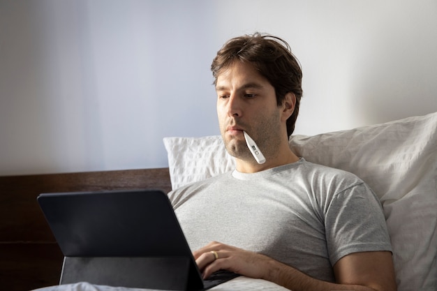 Man working in bed, doing home office because he is sick, with a thermometer in his mouth.