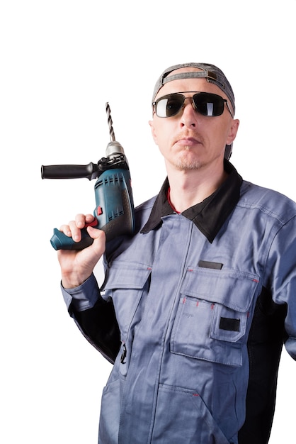 A man in working clothes, dark goggles and a cap, with an electric drill. A man of middle age, European appearance, in a working overalls. Isolated on white background