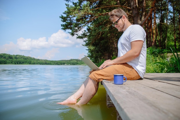 강물에 나무 부두 다리에 앉아있는 동안 노트북에서 작업하는 남자
