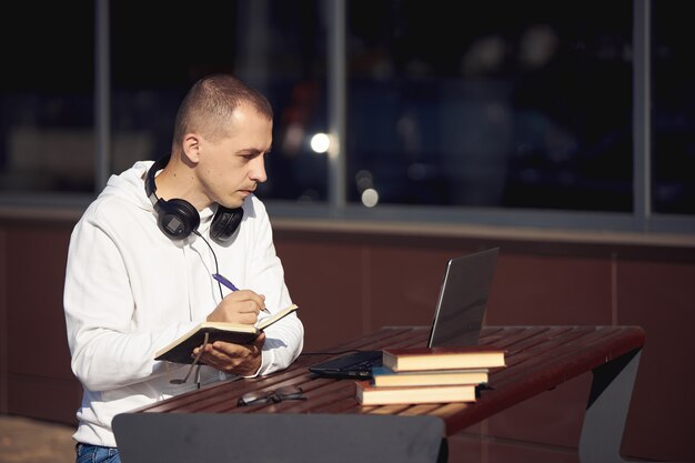 Man working on a laptop and writing in a notebook sitting on the street at a table. Social distancing during the coronavirus