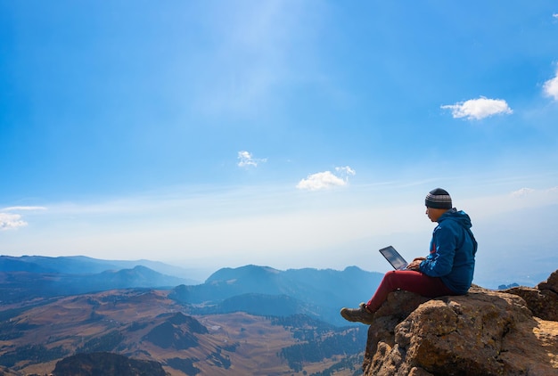 Man working on notebook sitting on cliff on top of the mountains