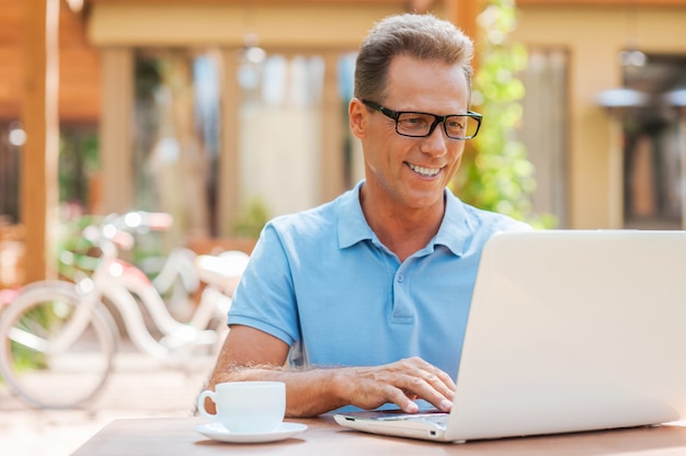 Man working outdoors. Cheerful mature man working at laptop and smiling while sitting at the table outdoors with house in the background