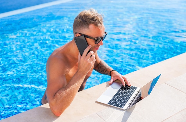 Man working on vacation speaking on phone and using laptop by the pool