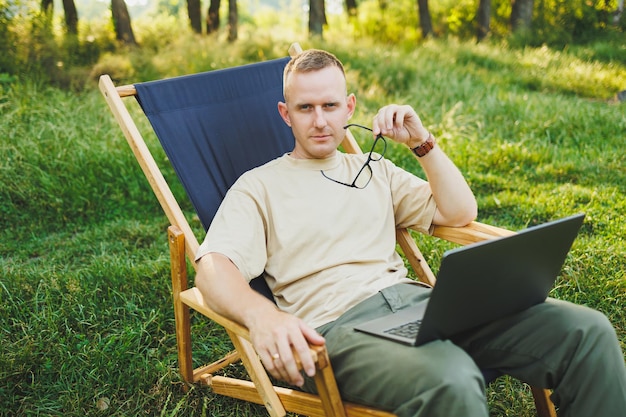 A man works on a laptop while sitting on a bench in a city park A young guy reads the news writes a message on his laptop Recreation in nature remote work on vacation