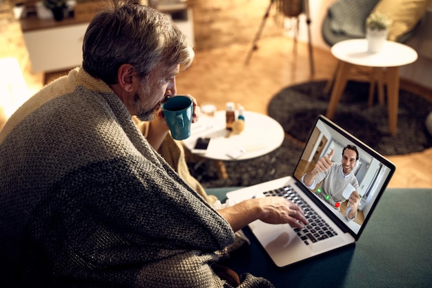 Man wrapped in blanket drinking tea while having video call\
with a colleague in the evening at home