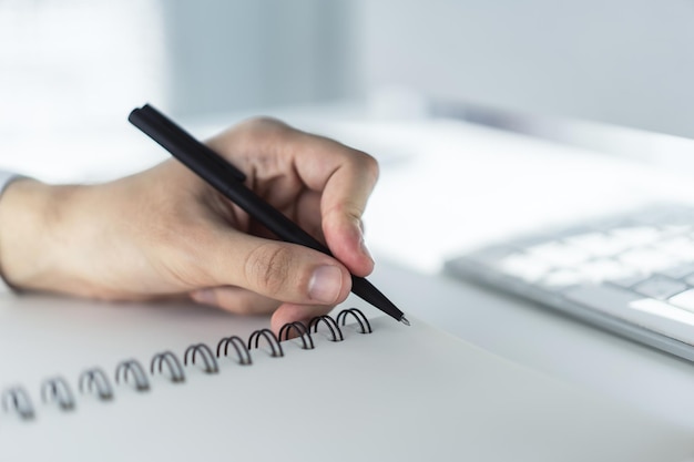 Man writes with a pen in notepad on computer keyboard in a sunny office business and education concept Close up