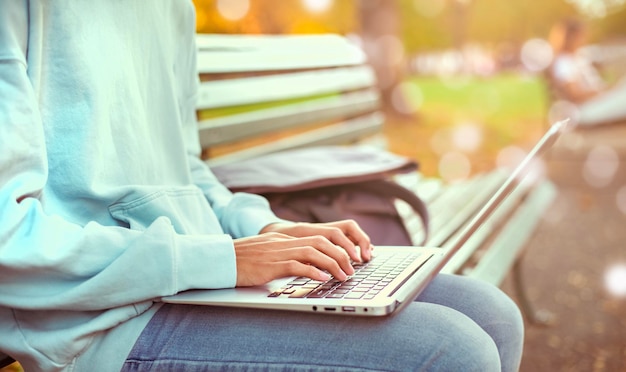 Man writing notebook with laptop in the park vintage style Slim hipster man in jeans using a notebook Freelancers communication Teenage student boy browses the Internet working outside the home