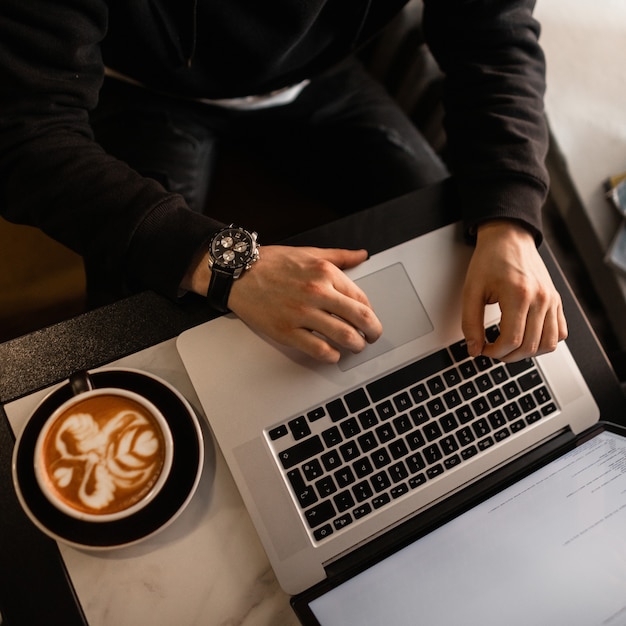 L'uomo con un orologio di lusso in una felpa con cappuccio nera si siede in un bar, beve caffè e lavora su un laptop al chiuso